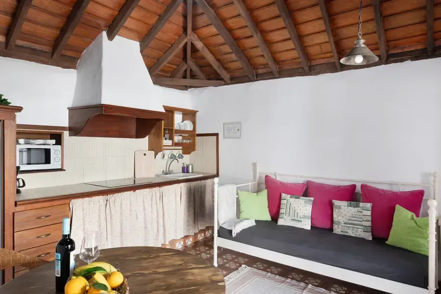 Cocina y zona de estar con vigas de madera, paredes blancas y almohadas de colores.