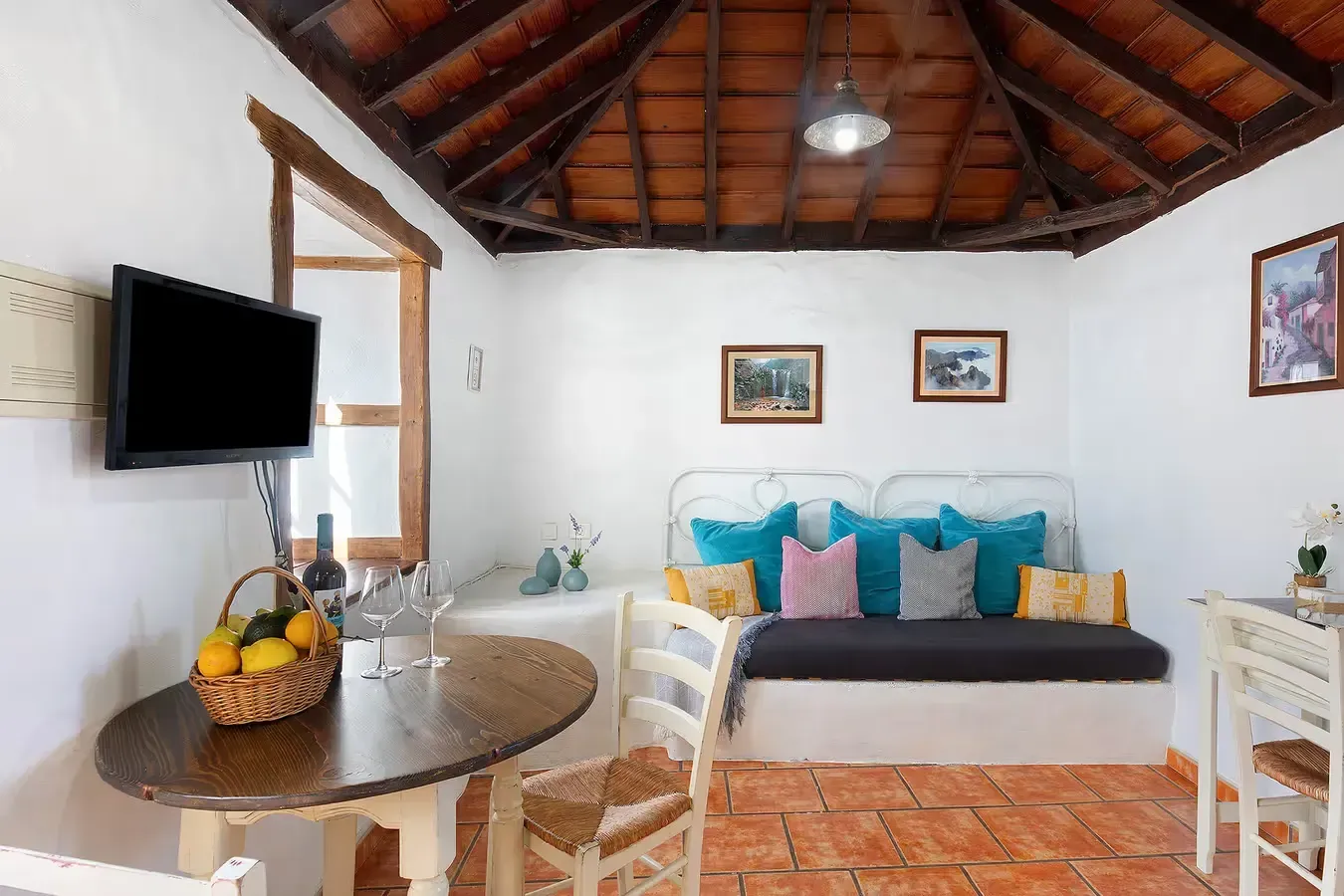 Acogedora sala de estar con sofá, mesa redonda, televisor y cuadros. Paredes blancas, suelo de terracota y techo de madera.