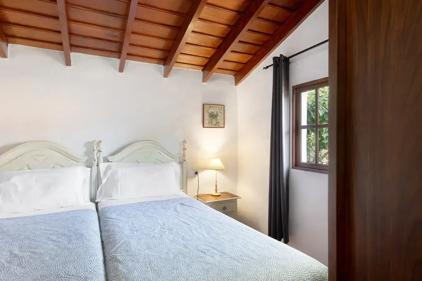 Dormitorio con dos camas individuales, paredes blancas, techo de madera, pequeña ventana con cortinas oscuras.