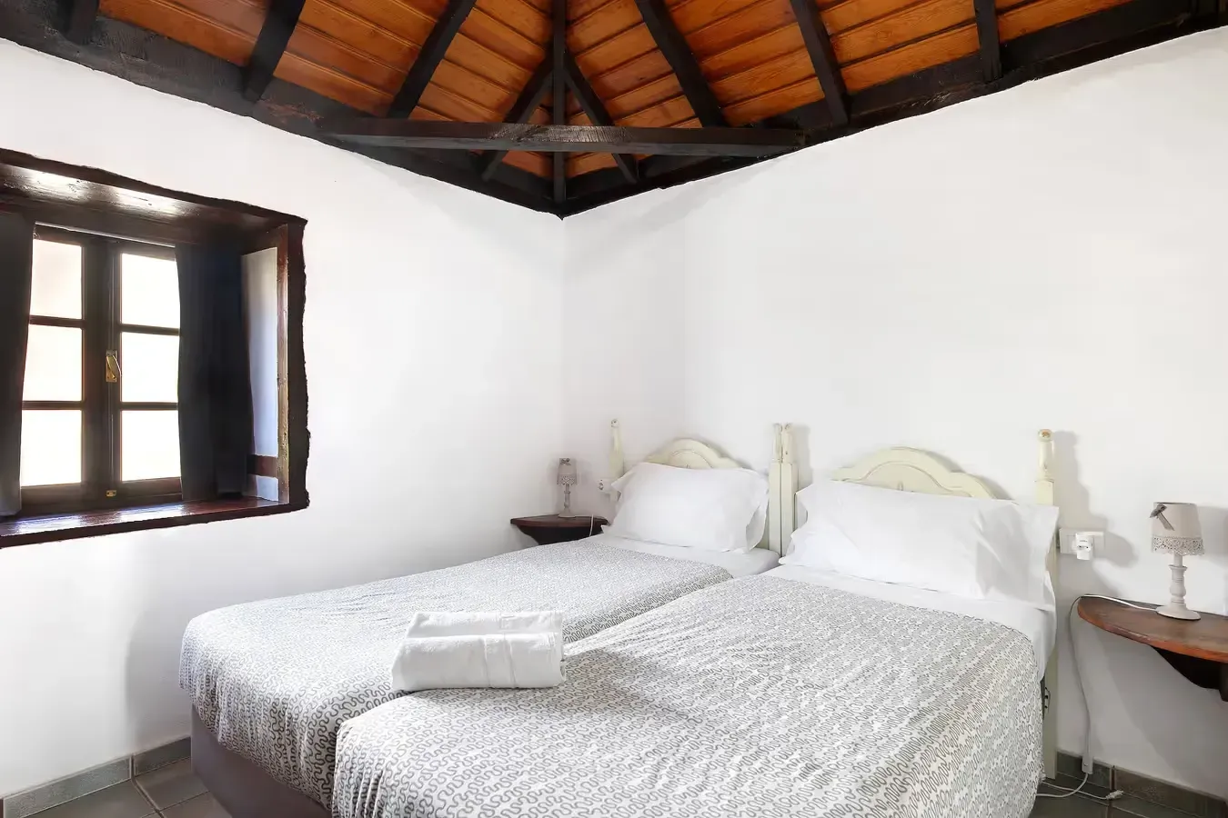 Dormitorio con dos camas individuales, paredes blancas, marco de ventana oscuro y techo de madera.