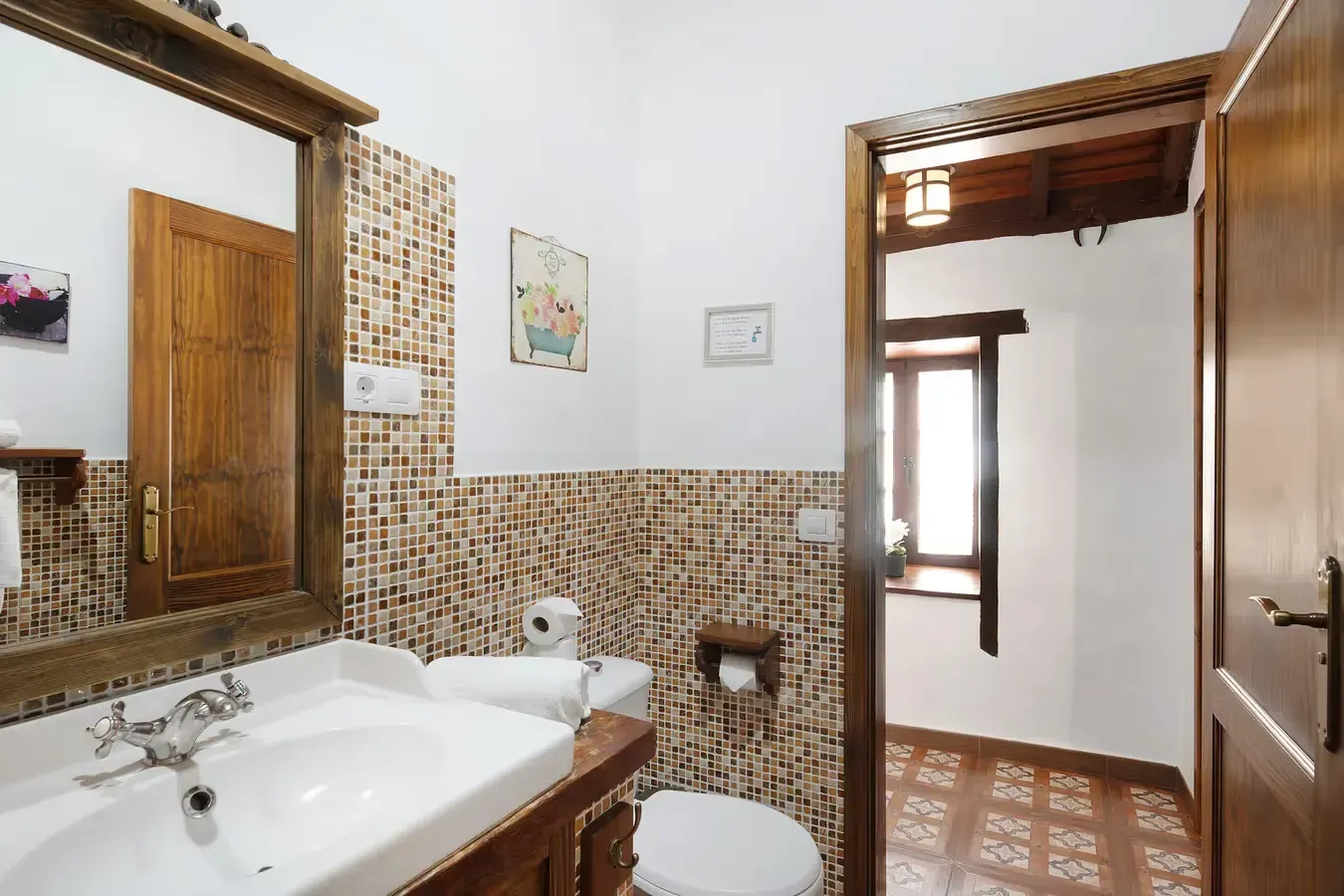 Baño con espejo con marco de madera, lavabo e inodoro, con puerta que da a una habitación con ventana.