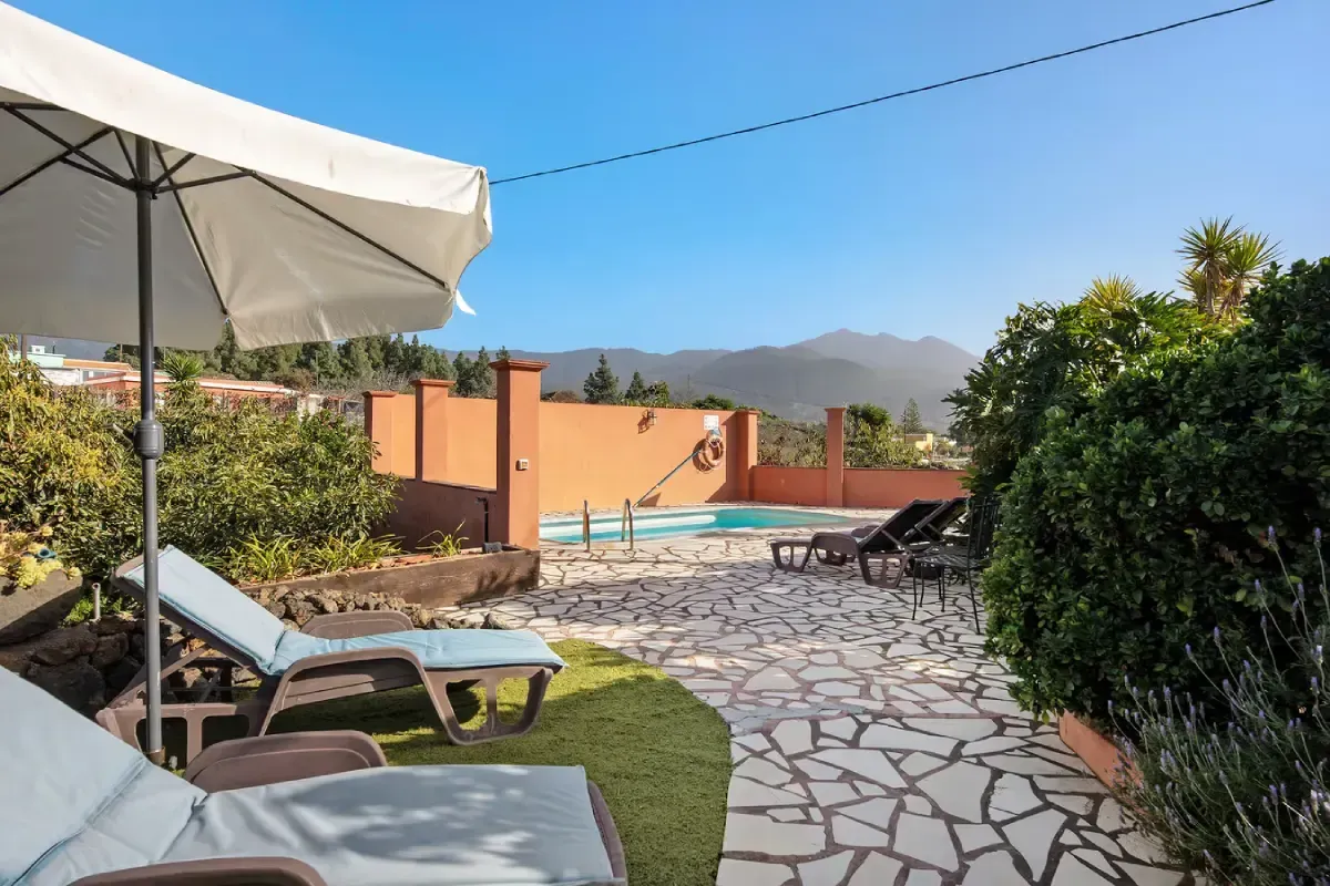 Área de piscina con tumbonas, sombrilla y vista a la montaña bajo un cielo azul.