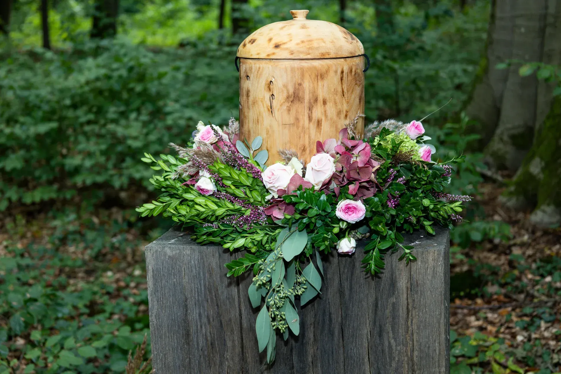 Urne aus Holz, auf einem Baumstumpf, umgeben von Blumen in einem Wald.