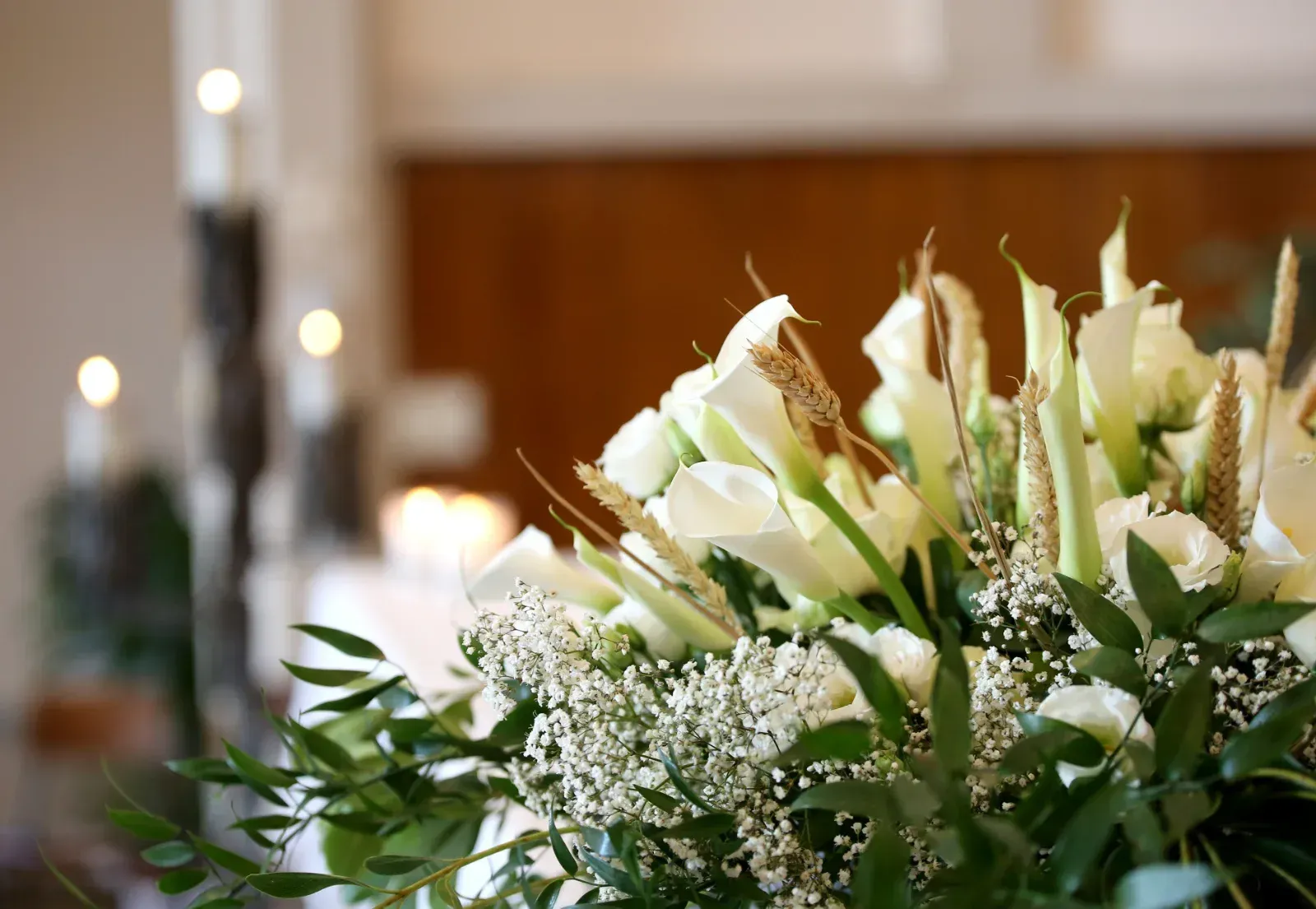 Weiße Calla-Lilien und Grünpflanzen in einem Strauß, mit Kerzen und Holzvertäfelung im Hintergrund.