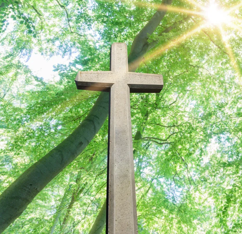 Betonkreuz vor dem Hintergrund leuchtend grüner Bäume und Sonnenlicht.