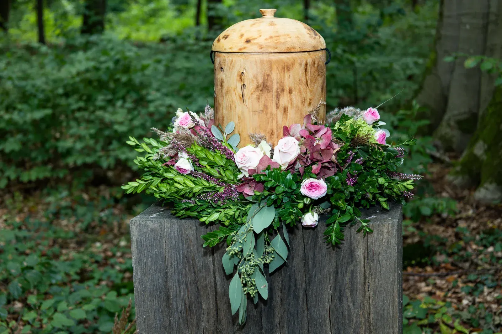 Holzurne mit Blumenkranz auf einem Baumstumpf in einem Wald.