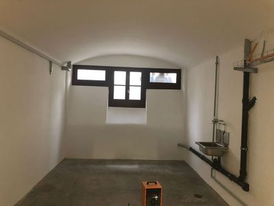Innenansicht eines kleinen Raums mit weißen Wänden, Fenster, Waschbecken und nacktem Betonboden.