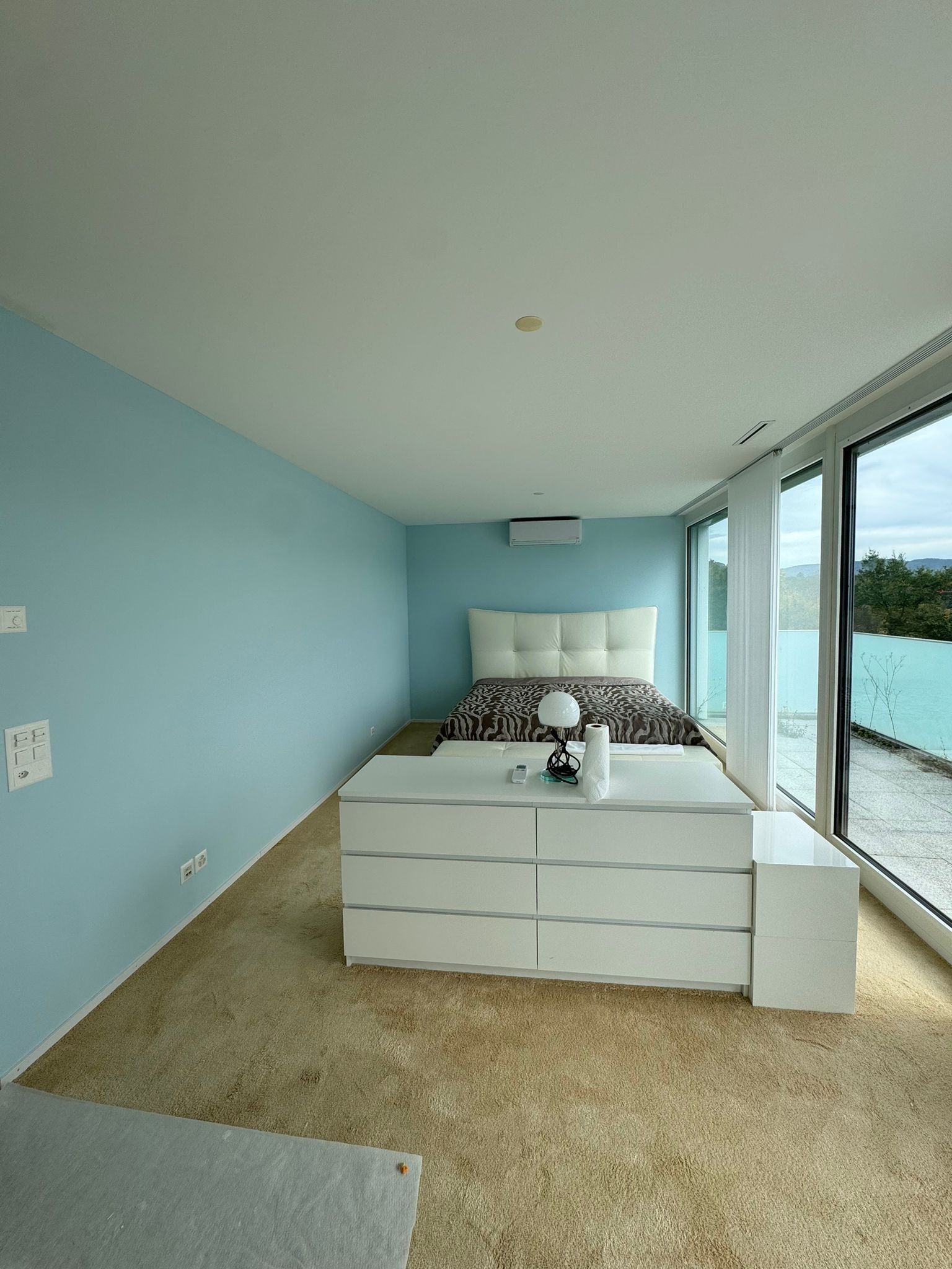 Schlafzimmer mit Bett, Kommode und großem Fenster mit Blick aufs Wasser. Hellblaue Wände, weiße Möbel.