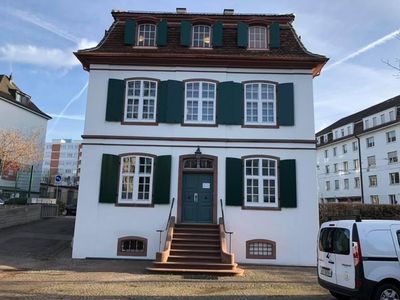 Weißes Gebäude mit grünen Fensterläden, braunem Dach. Stufen führen zu einer blauen Tür.