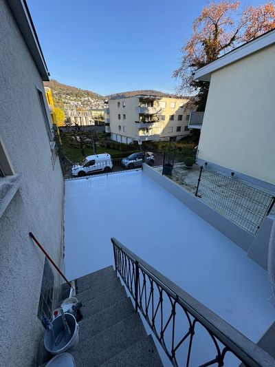 Außenansicht einer frisch gestrichenen grauen Terrasse. Im Hintergrund Gebäude und Bäume unter blauem Himmel.
