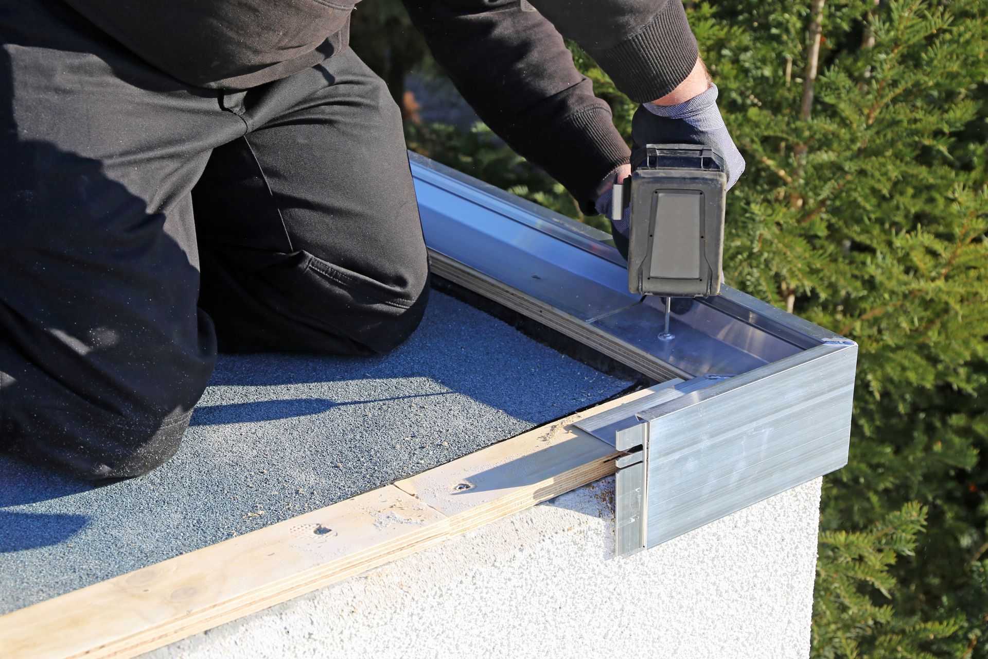 Un couvreur qui installe un solin métallique à l'aude d'une perceuse sur un toit plat.