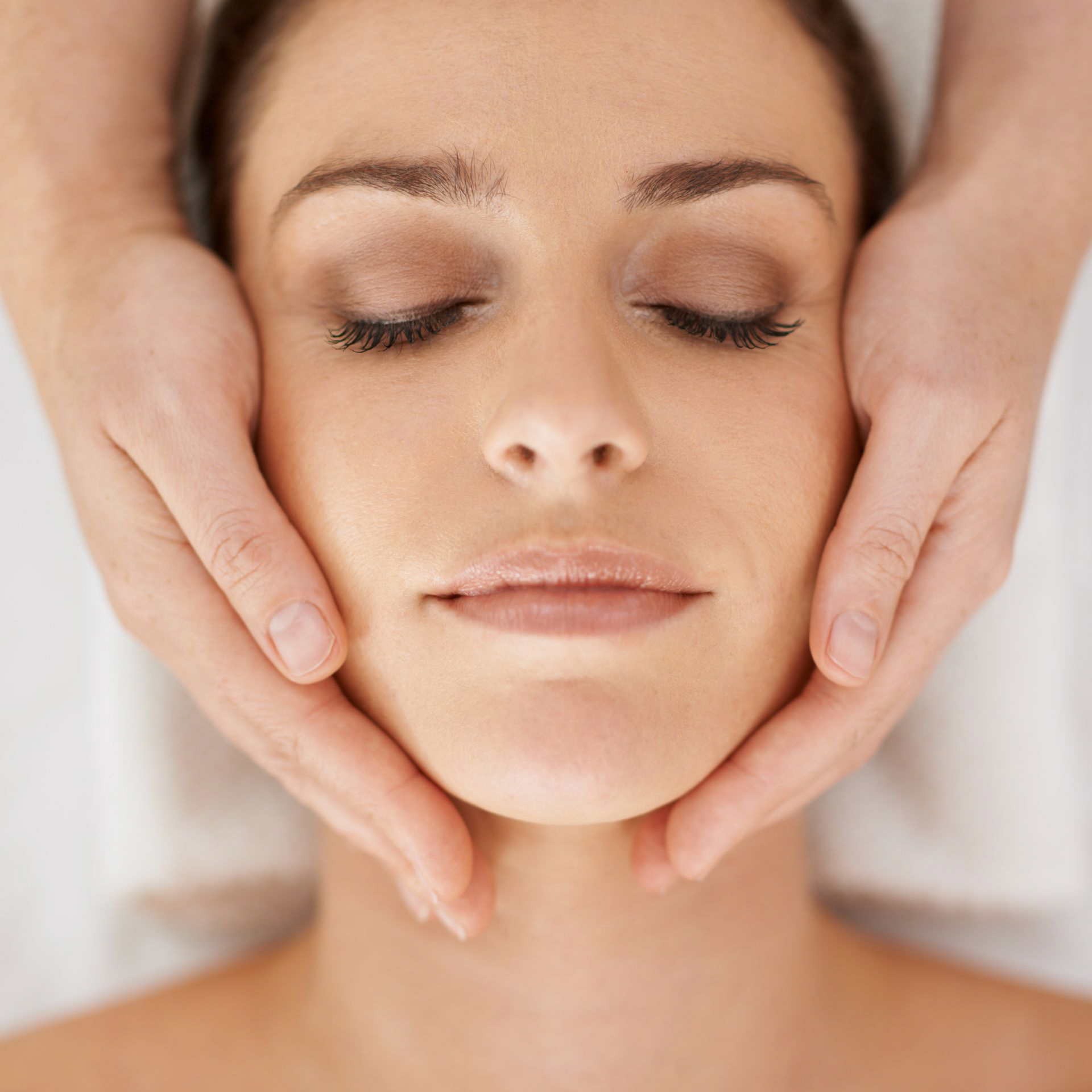 Visage d'une femme aux yeux clos, recevant un massage facial. Des mains caressent le visage ; lumière tamisée.