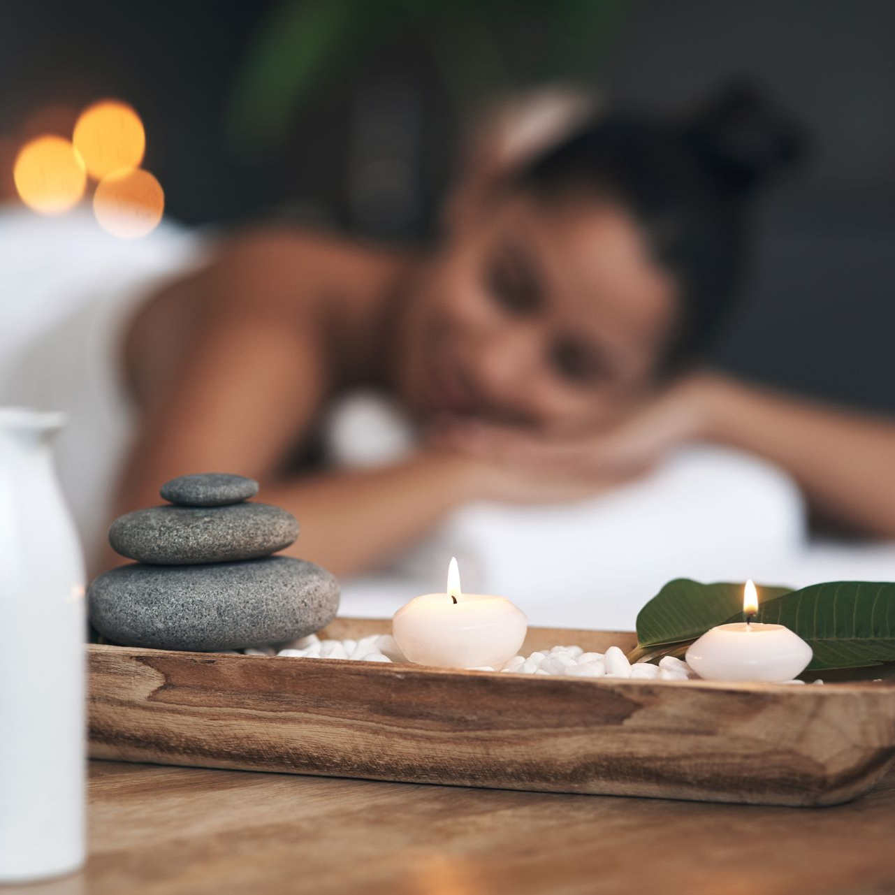 Des pierres, des bougies et une feuille sur un plateau en bois avec une femme recevant un massage en arrière-plan flou.