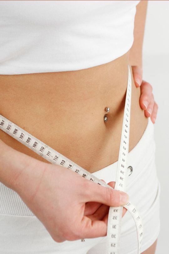 Personne mesurant sa taille avec un ruban à mesurer, piercing au nombril visible.