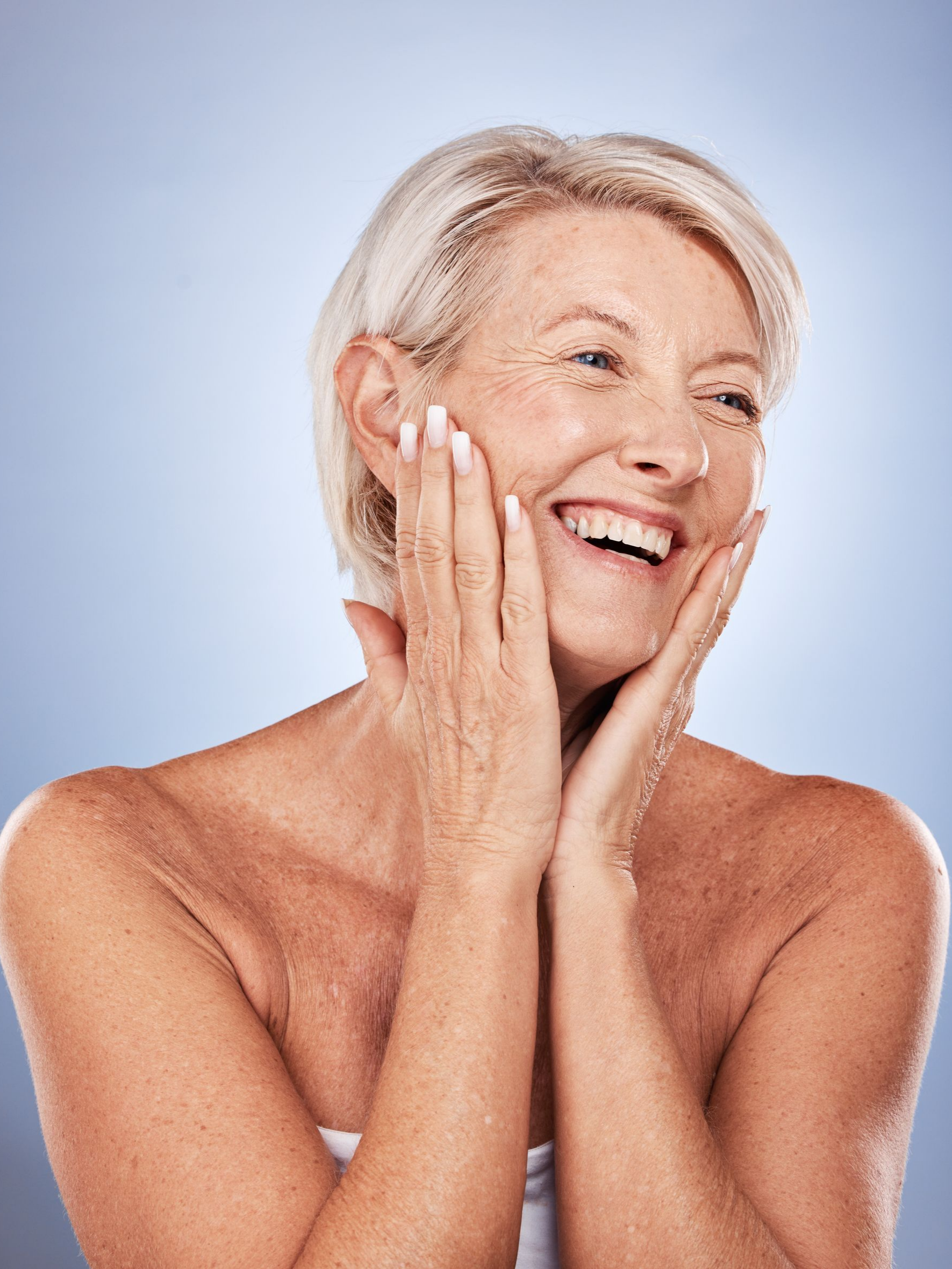 Femme aux cheveux courts et blancs, souriante et se touchant le visage avec les mains. Fond bleu.