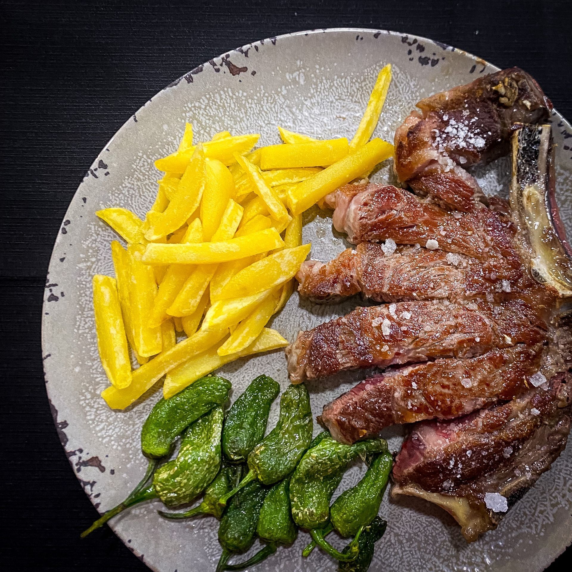 Plato con filete en rodajas, patatas fritas amarillas y pimientos verdes sobre una mesa oscura.