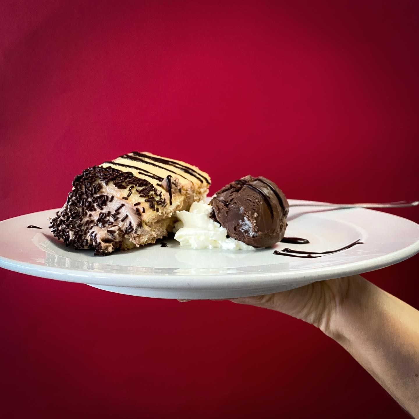 Postre en un plato blanco: pastel bañado en chocolate y una bola de helado sobre un fondo rojo.