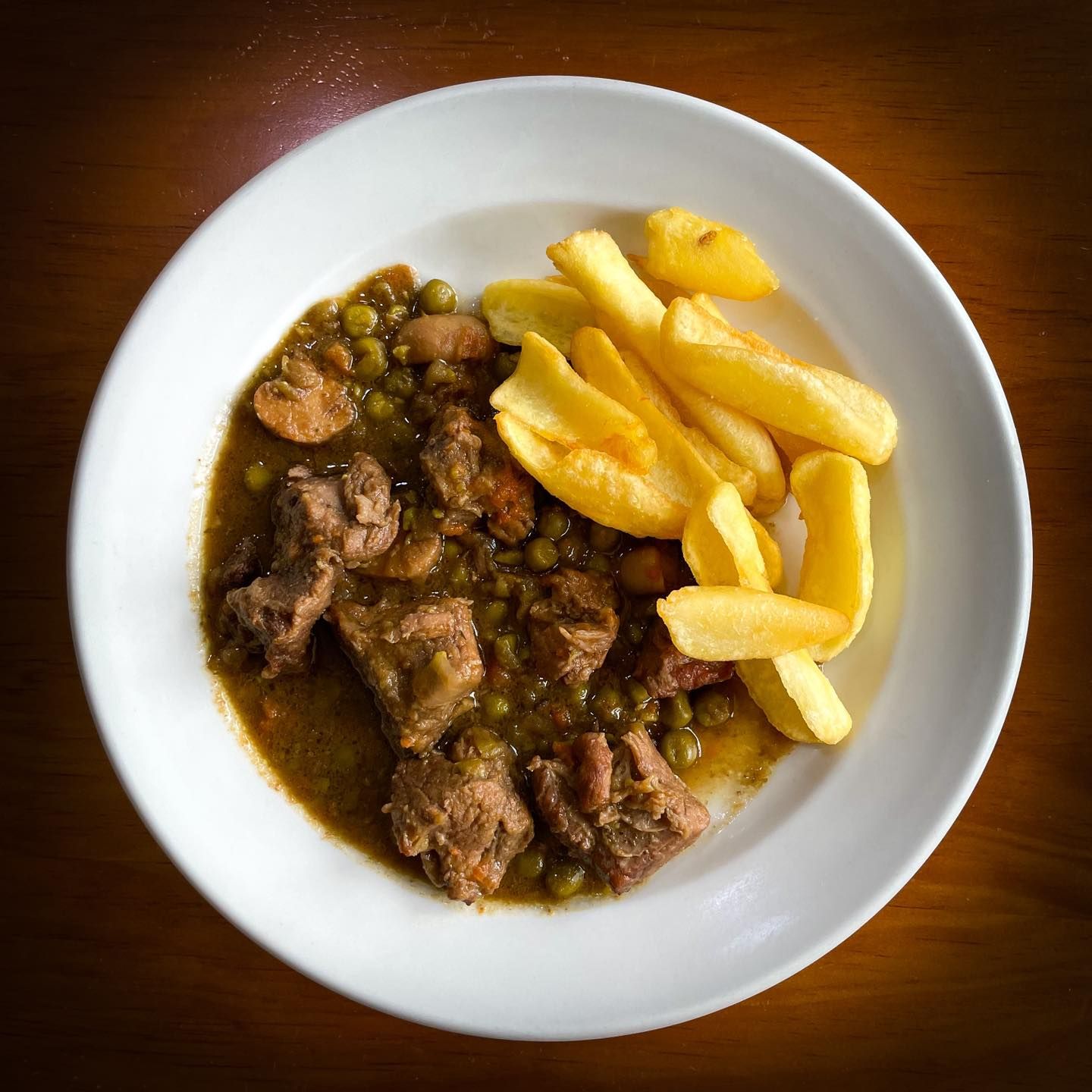 Un bol de estofado de ternera con guisantes y patatas fritas gruesas en un plato blanco.