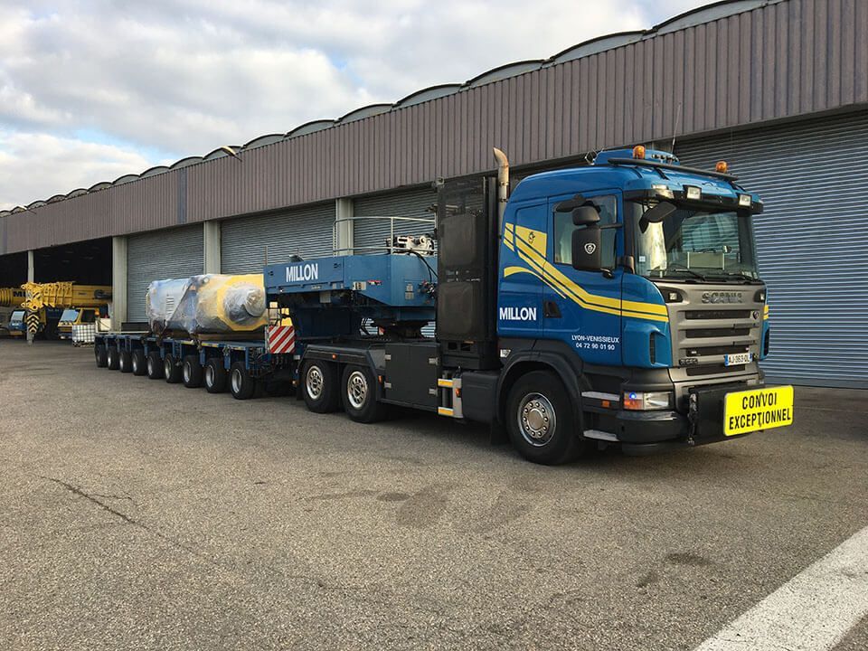 Un camion Scania bleu, poids lourd, transporte un gros composant industriel emballé sur une remorque à plusieurs essieux, sur un parking.