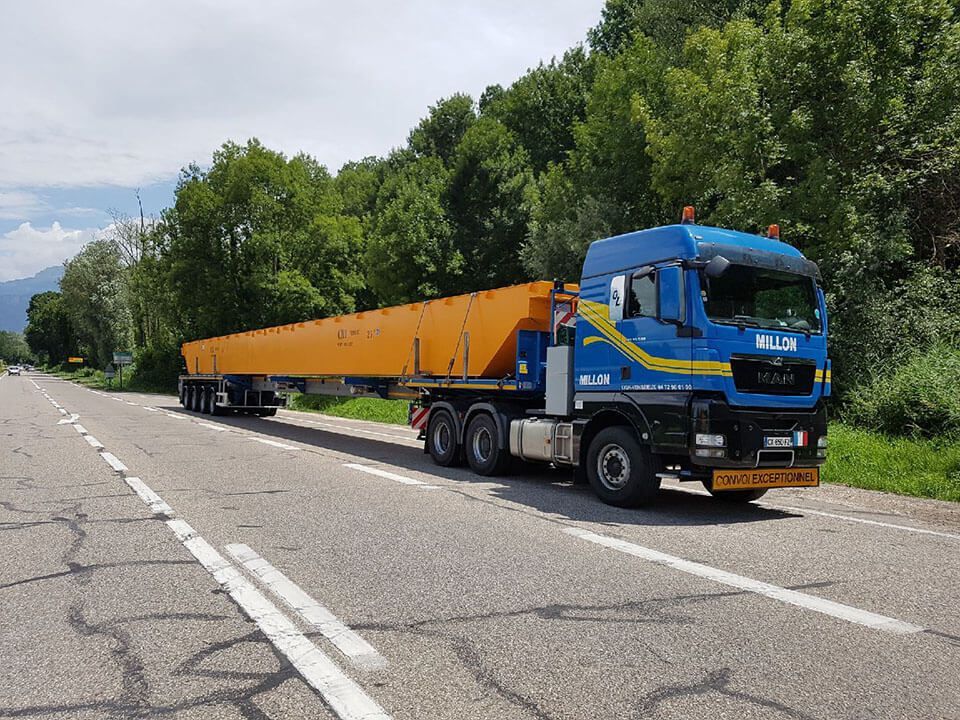 Un semi-remorque bleu tirant une longue remorque plate orange chargée d'une énorme poutre métallique sur une route goudronnée.