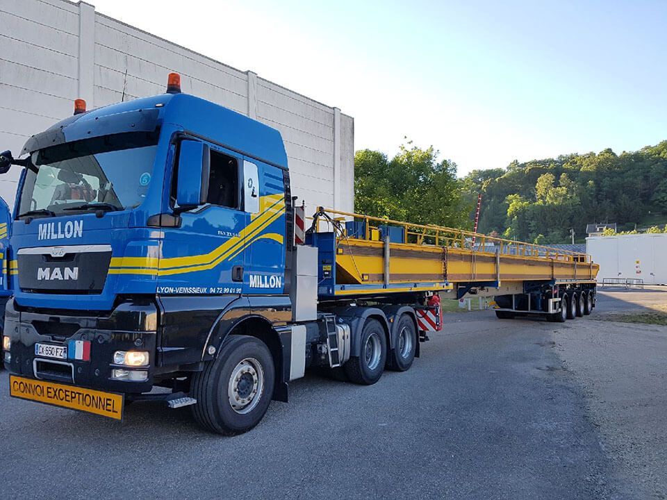 Un camion MAN bleu, poids lourd, avec une remorque plateau jaune, est stationné sur un parking asphalté contre un mur blanc.