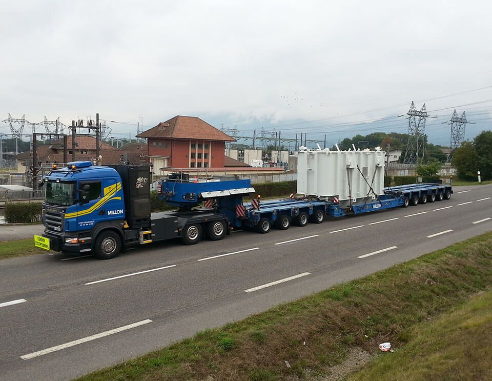 Un camion bleu poids lourd transportant un gros transformateur industriel blanc sur une remorque à plusieurs essieux, le long d'une route asphaltée.