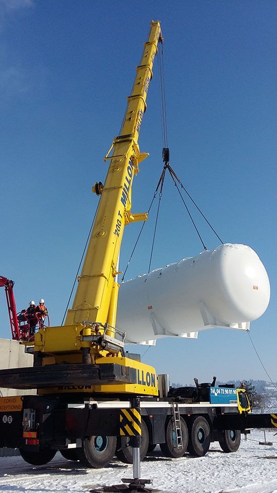 Une grue mobile jaune soulève un grand réservoir de stockage horizontal blanc sur fond de ciel bleu clair, au-dessus d'un sol enneigé.