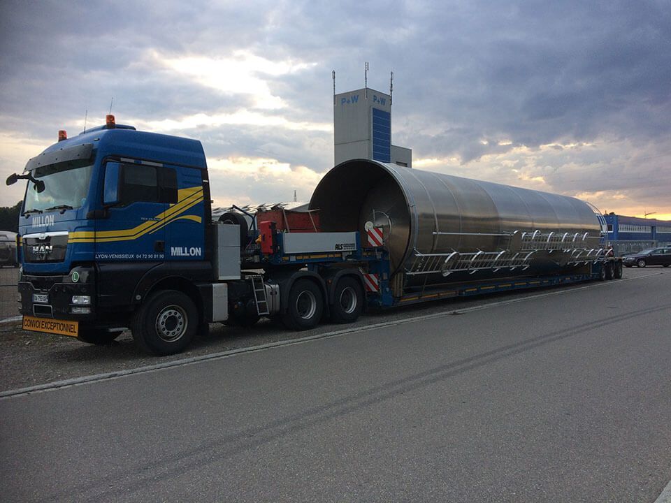 Un semi-remorque bleu tire une longue cuve métallique industrielle cylindrique sur une remorque, sur un terrain vague.