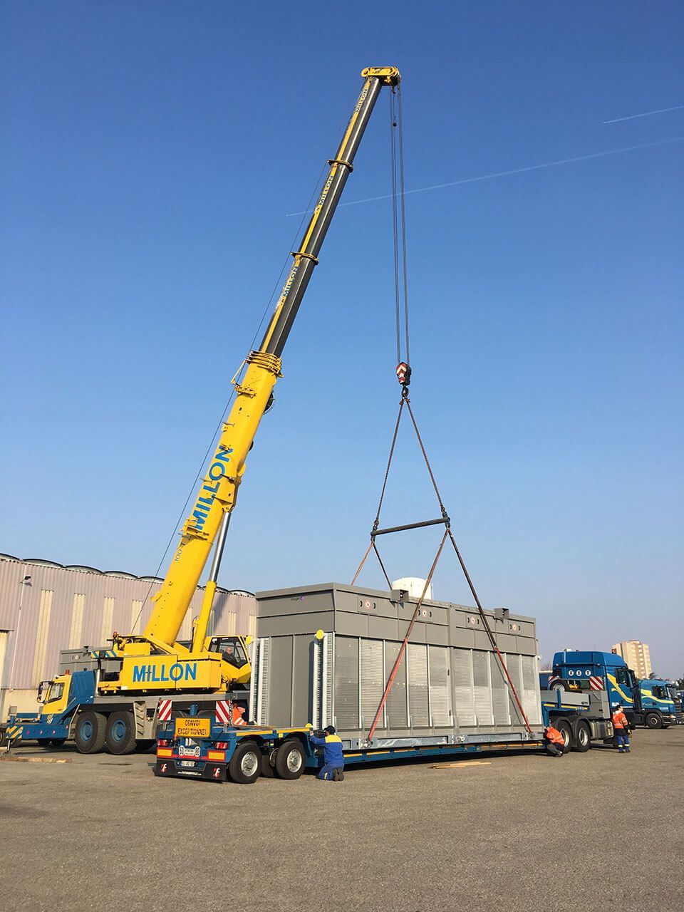 Une grue jaune soulève une grande unité industrielle d'un camion plateau sur un terrain goudronné et dégagé, sous un ciel bleu azur.