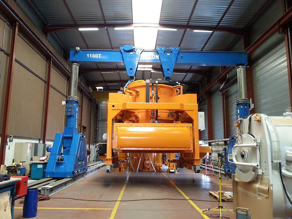 Une grande grue industrielle soulève une lourde pièce de machine jaune à l'intérieur d'un vaste entrepôt.