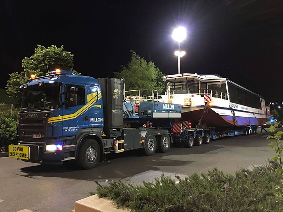 Un camion Scania bleu transportant de nuit un grand catamaran blanc et rouge sous un puissant projecteur.