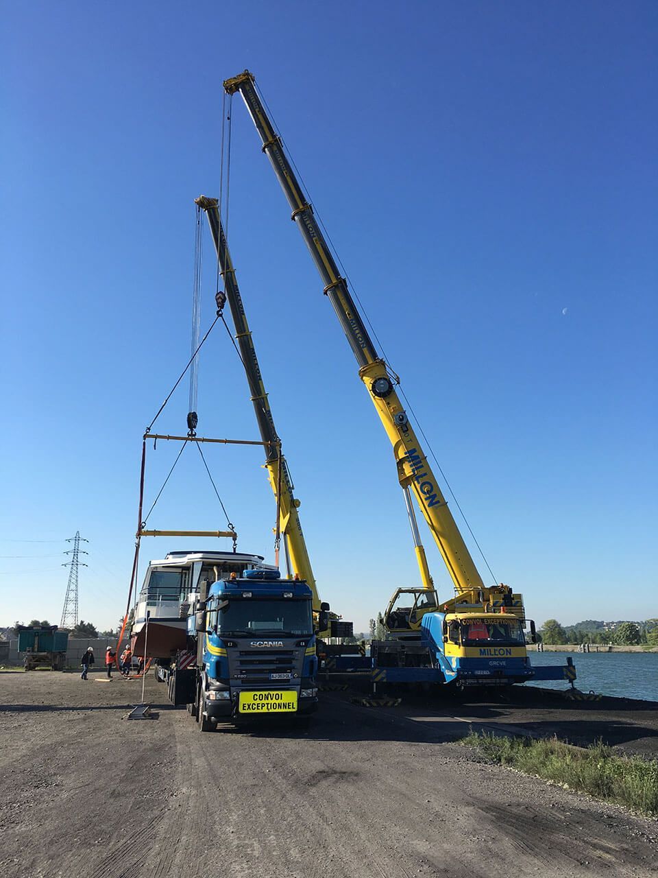 Deux grues jaunes travaillent de concert pour soulever un bateau et le déposer sur un camion de transport bleu stationné sur un terrain de gravier près d'un plan d'eau.