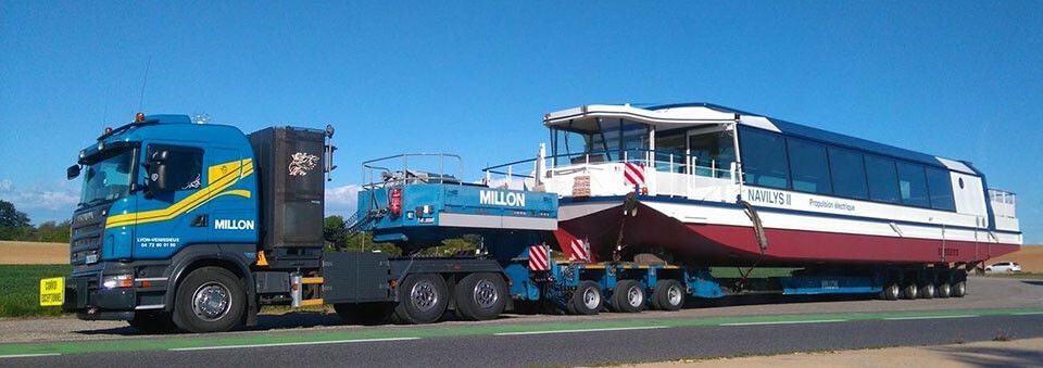 Un camion bleu poids lourd transporte un grand bateau de passagers sur une remorque à plateau, le long d'une route sous un ciel bleu dégagé.