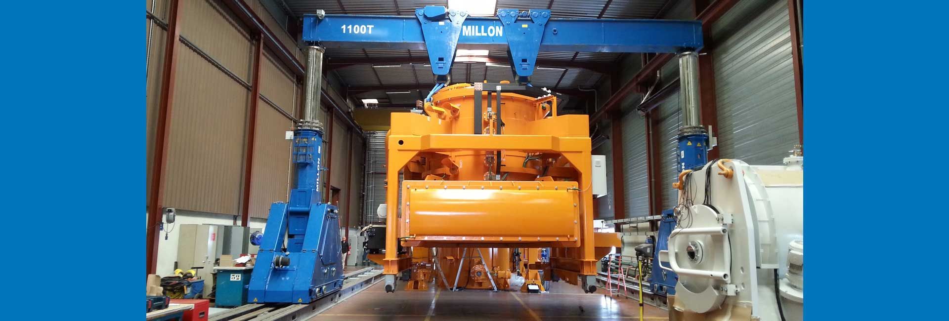 Une grande grue portique bleue soulève une lourde machine industrielle orange à l'intérieur d'un entrepôt ou d'une usine.