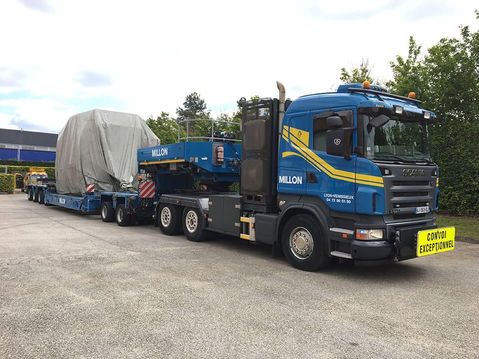 Un camion de transport Milon bleu transporte un objet volumineux recouvert d'une bâche grise sur une remorque à plateau, sur un parking goudronné.