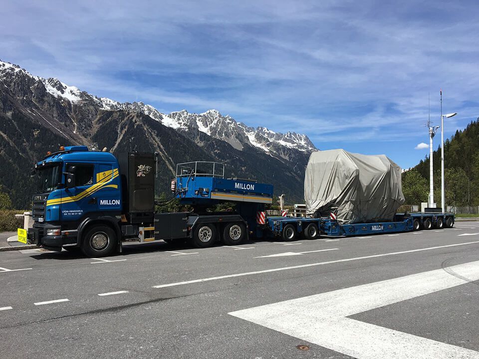 Un semi-remorque Scania bleu transportant une importante cargaison industrielle recouverte d'une bâche sur une remorque à plateau près de montagnes enneigées.