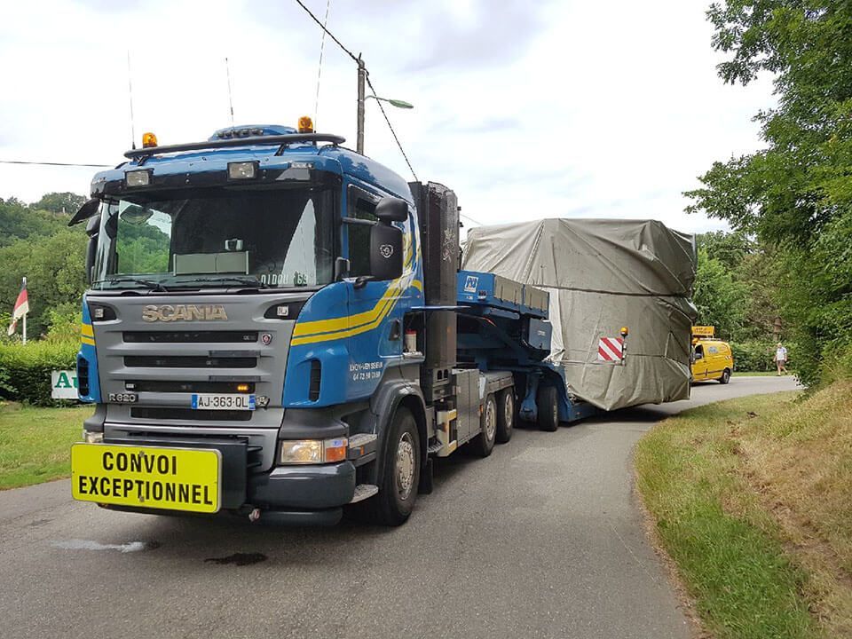 Un camion Scania bleu, arborant un panneau jaune CONVOI EXCEPTIONNEL , tire un chargement volumineux et bâché sur une route étroite bordée d'arbres.