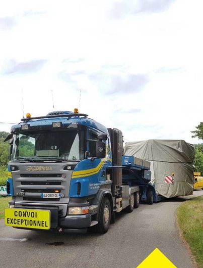 Un semi-remorque Scania bleu transportant une importante cargaison industrielle recouverte d'une bâche sur une route, portant la mention « CONVOI EXCEPTIONNEL ».