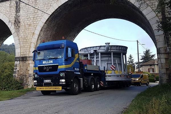 Un semi-remorque MAN bleu transporte une grande cuve industrielle circulaire sous un pont en pierre en arc.