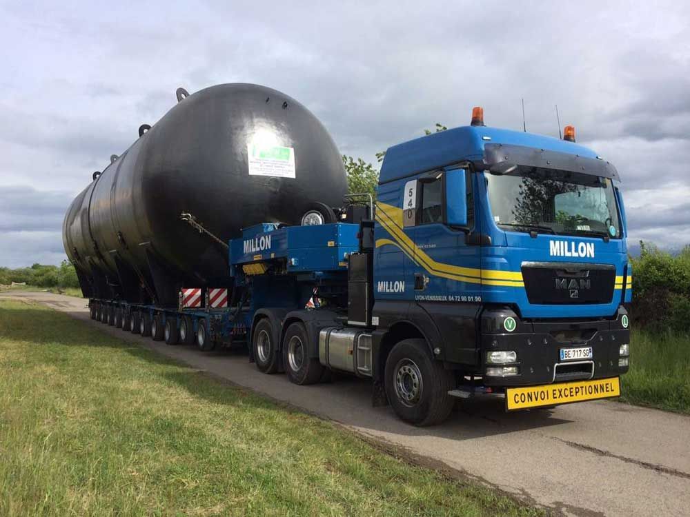 Un semi-remorque MAN bleu transportant une cuve industrielle cylindrique noire surdimensionnée sur une remorque à plusieurs essieux, le long d'une route asphaltée.