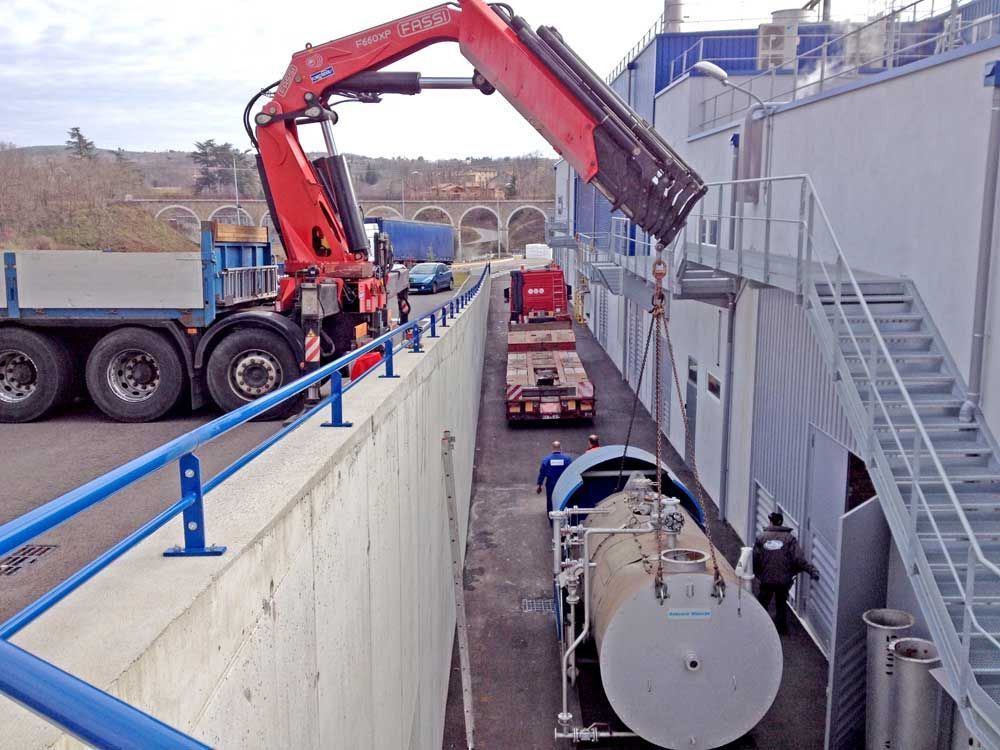 Une grue rouge montée sur camion descend un grand réservoir cylindrique d'eau grise dans un espace étroit entre un mur de soutènement et un bâtiment.