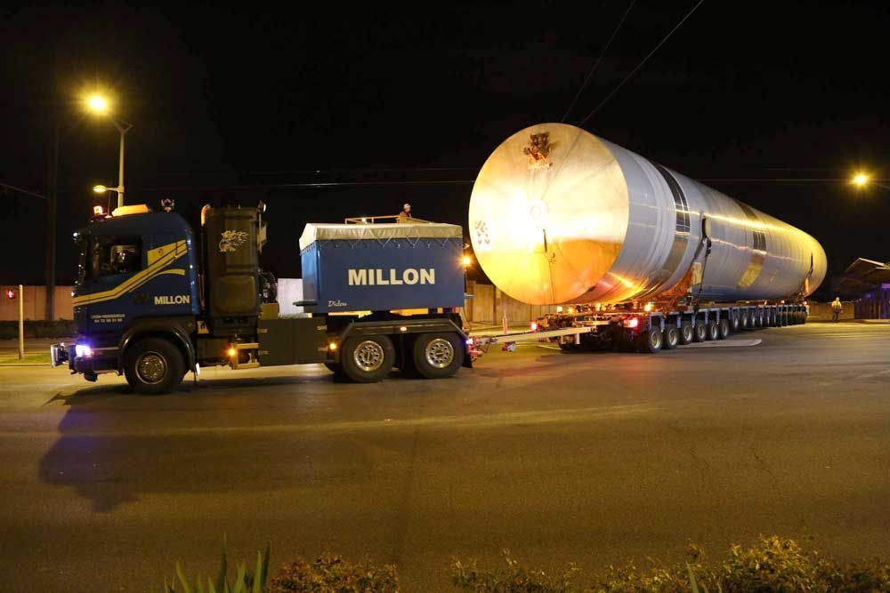Un camion bleu Millon transporte un grand réservoir cylindrique sur une remorque à plusieurs essieux, de nuit, sur une route.