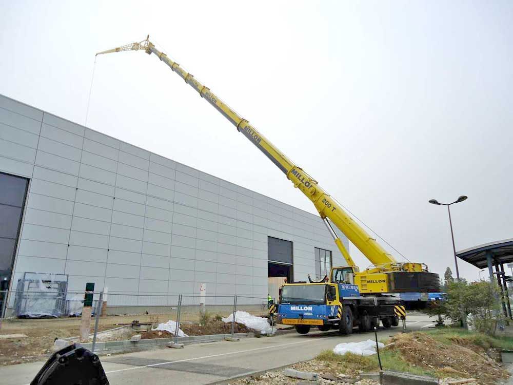 Une grue mobile jaune à flèche télescopique déployée est stationnée devant un bâtiment industriel gris.