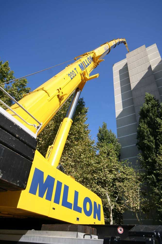 Une grue jaune Millon déploie sa flèche vers un grand bâtiment en béton gris sous un ciel bleu azur.