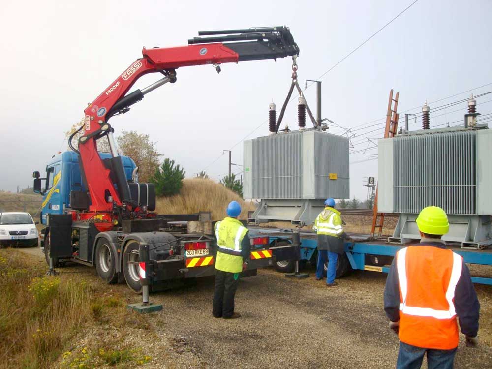 Un camion-grue soulève un gros transformateur électrique tandis que trois ouvriers en gilets haute visibilité supervisent l'opération sur un terrain vague.