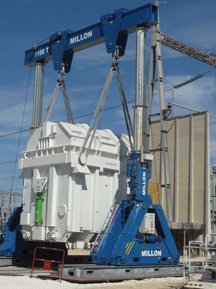 Une grande grue portique bleue soulève un transformateur industriel blanc dans un poste de transformation électrique extérieur.