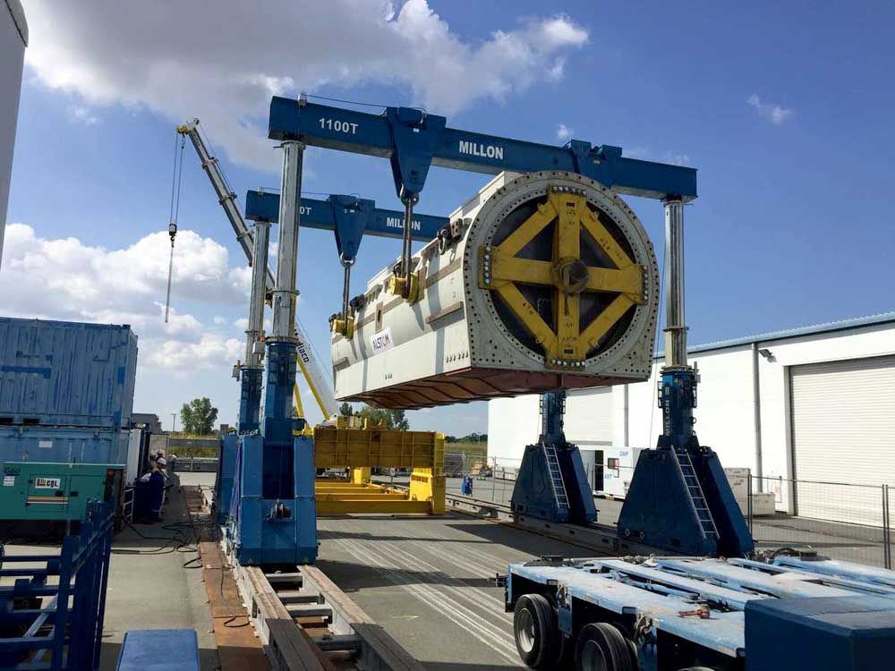 À l'extérieur, une grue à portique bleue soulève un grand composant industriel blanc muni d'une entretoise jaune en forme de croix.