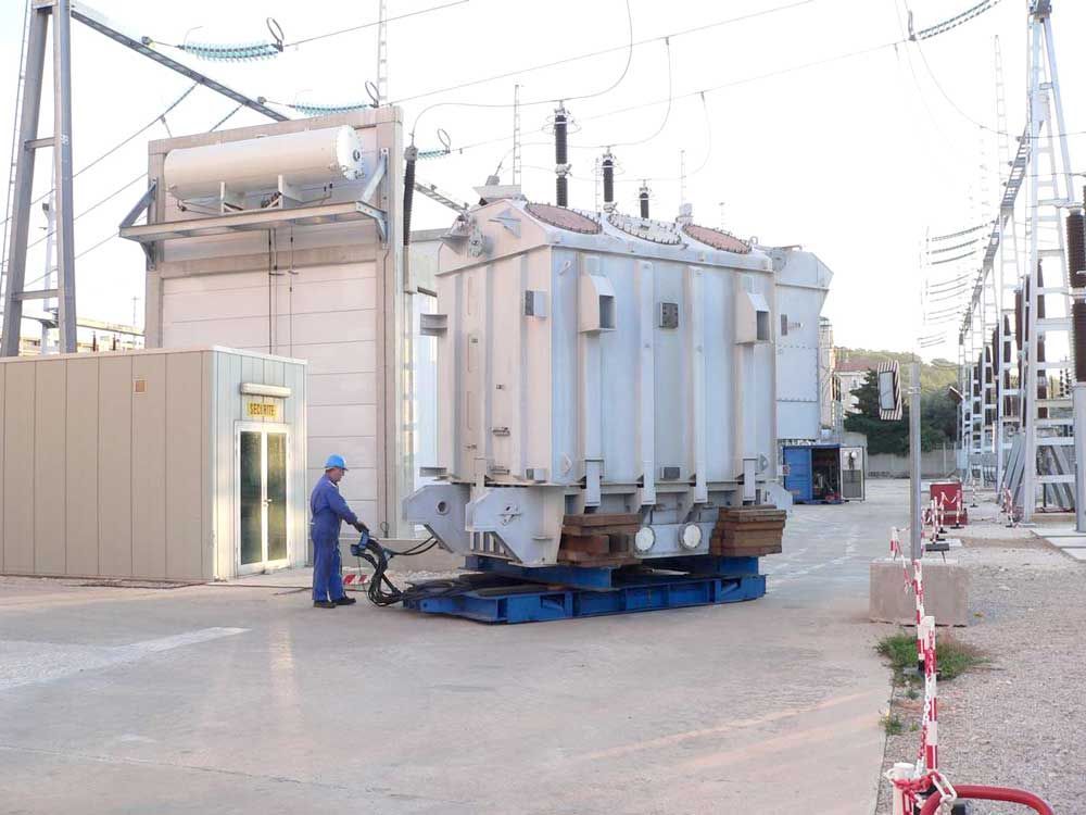 Un ouvrier en uniforme bleu manœuvre un gros transformateur électrique sur une plateforme robuste dans une sous-station électrique.
