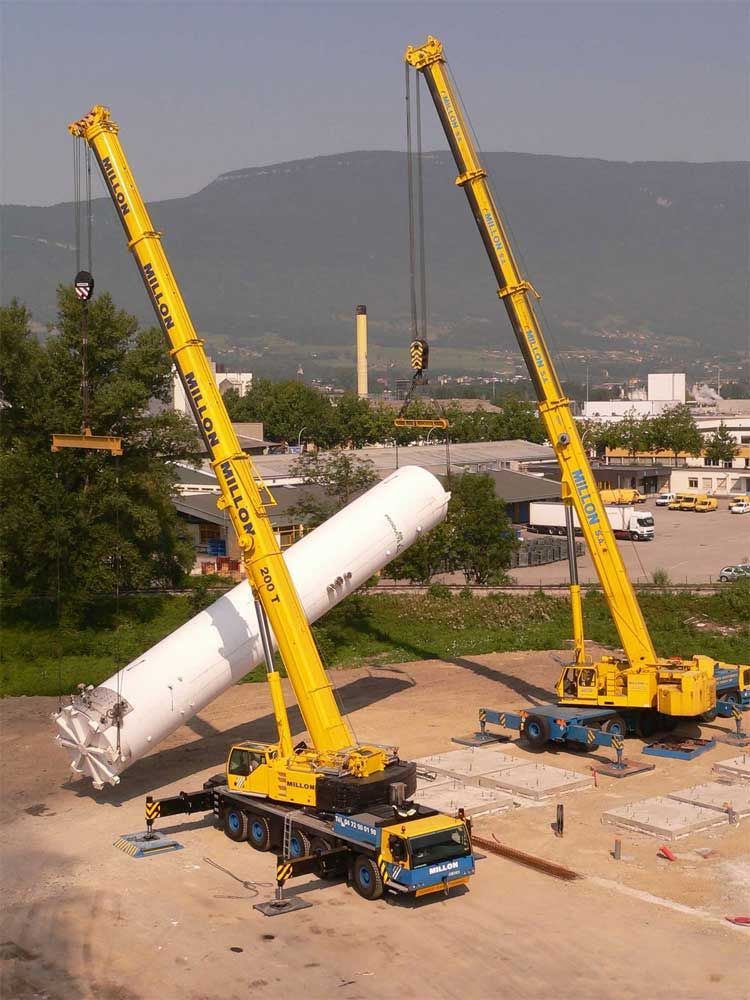 Deux grandes grues mobiles jaunes travaillent en tandem pour soulever un lourd objet cylindrique blanc sur un chantier de construction en extérieur.