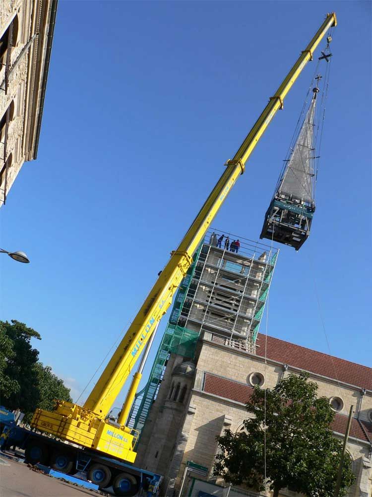 Sous un ciel bleu limpide, une grue de chantier jaune soulève la flèche d'un clocher au-dessus d'un bâtiment en pierre recouvert d'échafaudages.